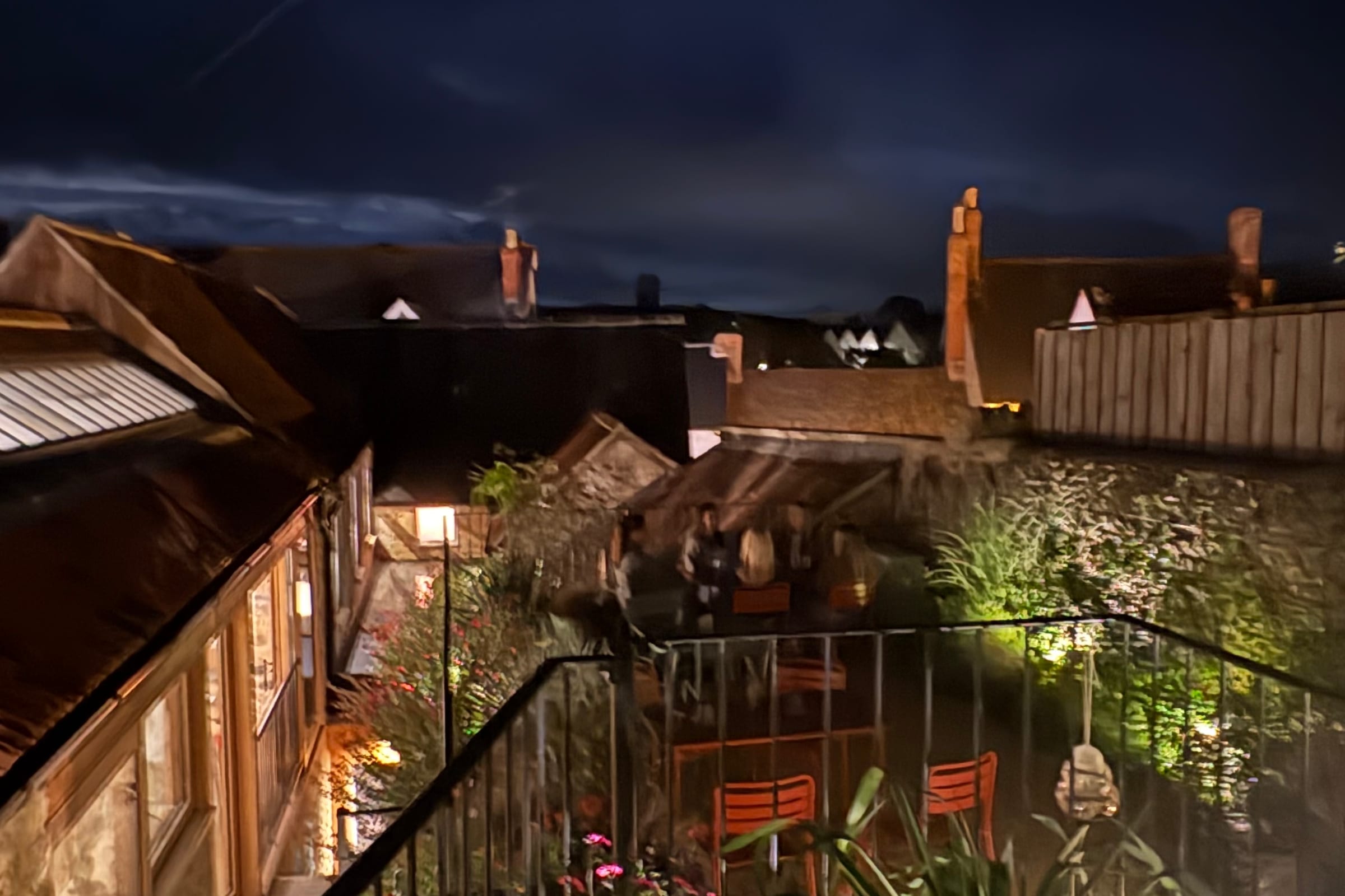 Looking down from the top of The Prickly Pear's garden in Bruton at night: lit flower beds and orange chairs behind railings, a few diners at the far tables, and the town's rooftops and chimneys under a deep blue sky