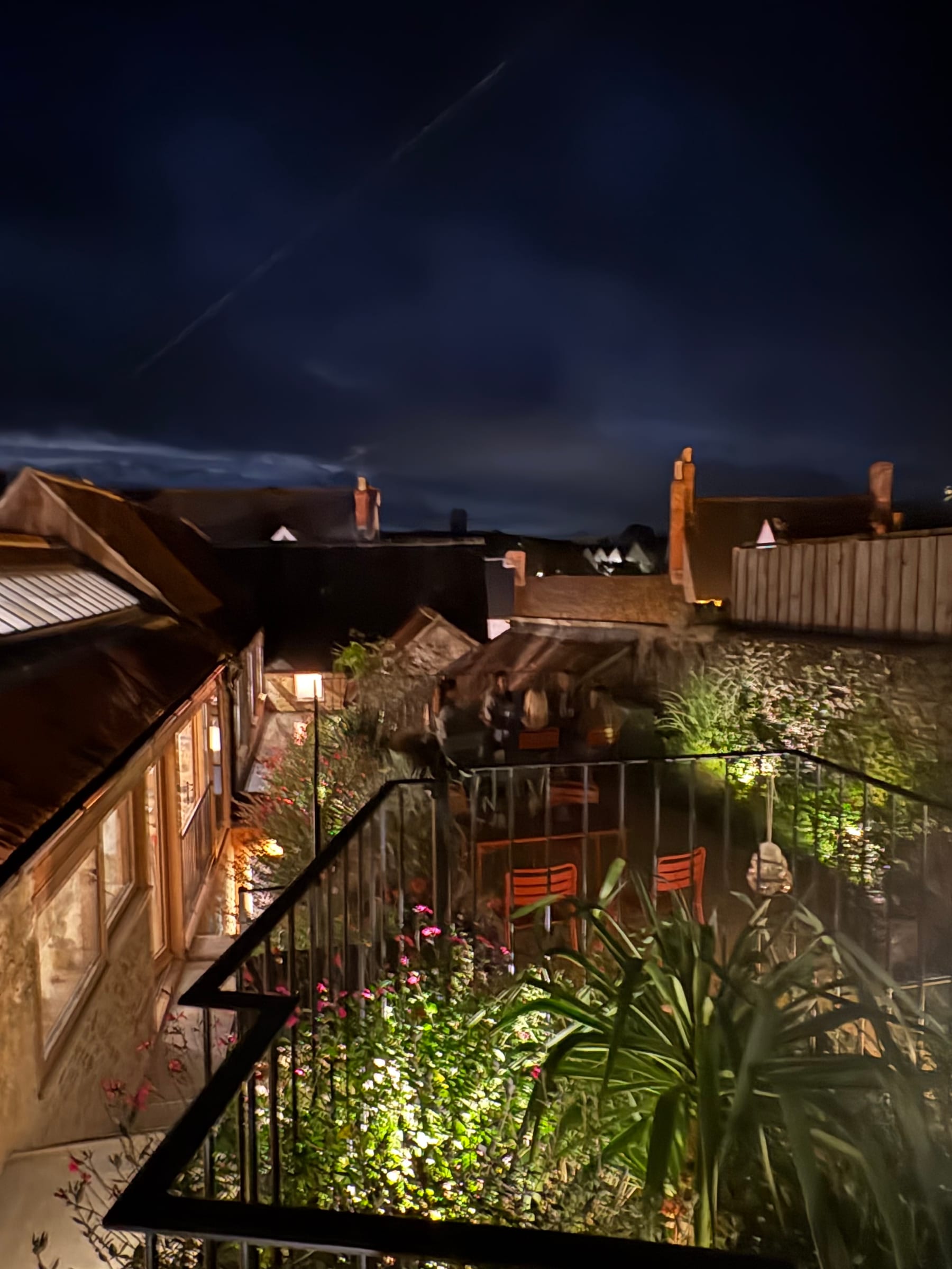 Looking down from the top of The Prickly Pear's garden in Bruton at night: lit flower beds and orange chairs behind railings, a few diners at the far tables, and the town's rooftops and chimneys under a deep blue sky crossed by a vapour trail